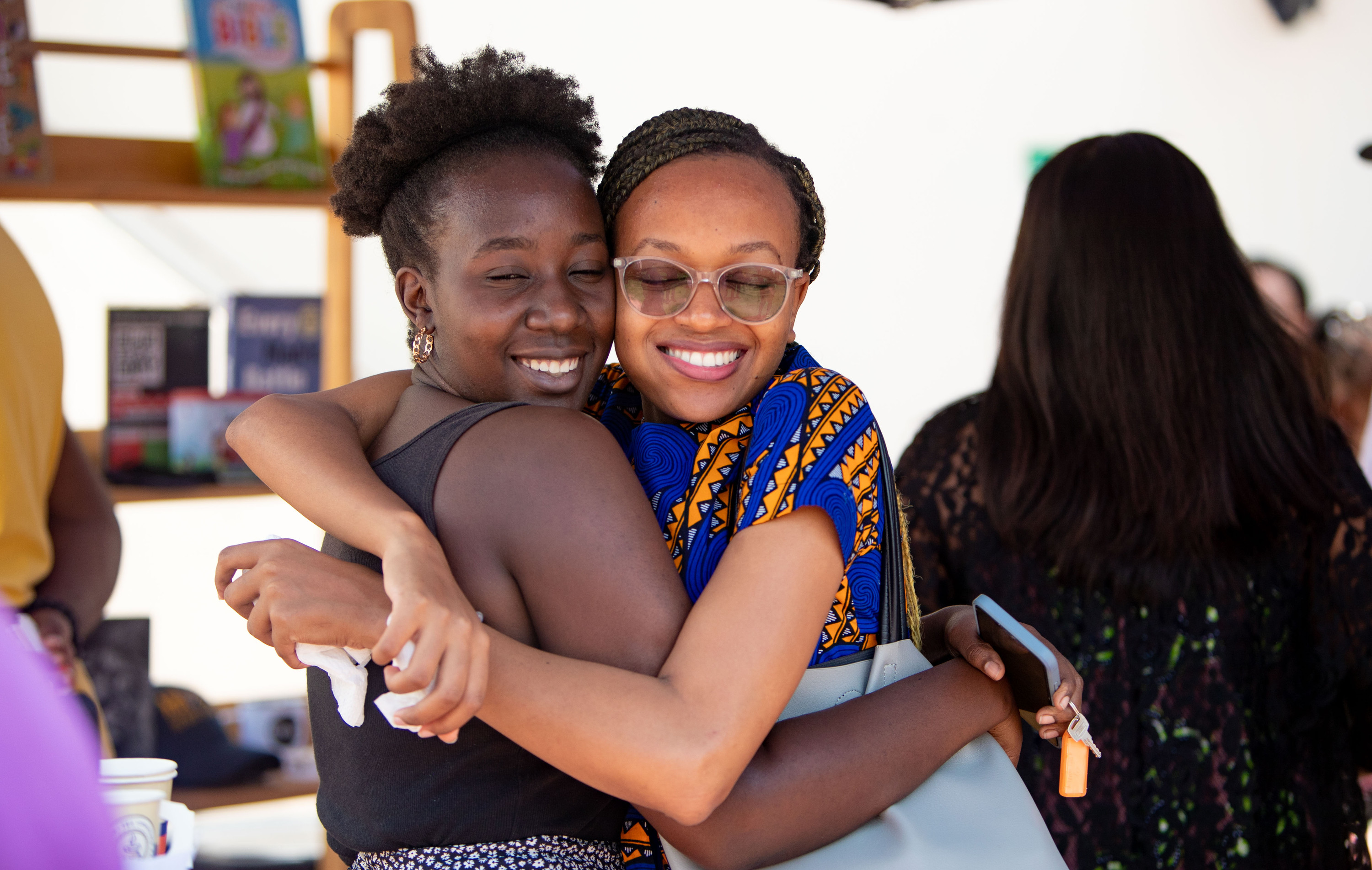 Two women hug and smile beside a book stall